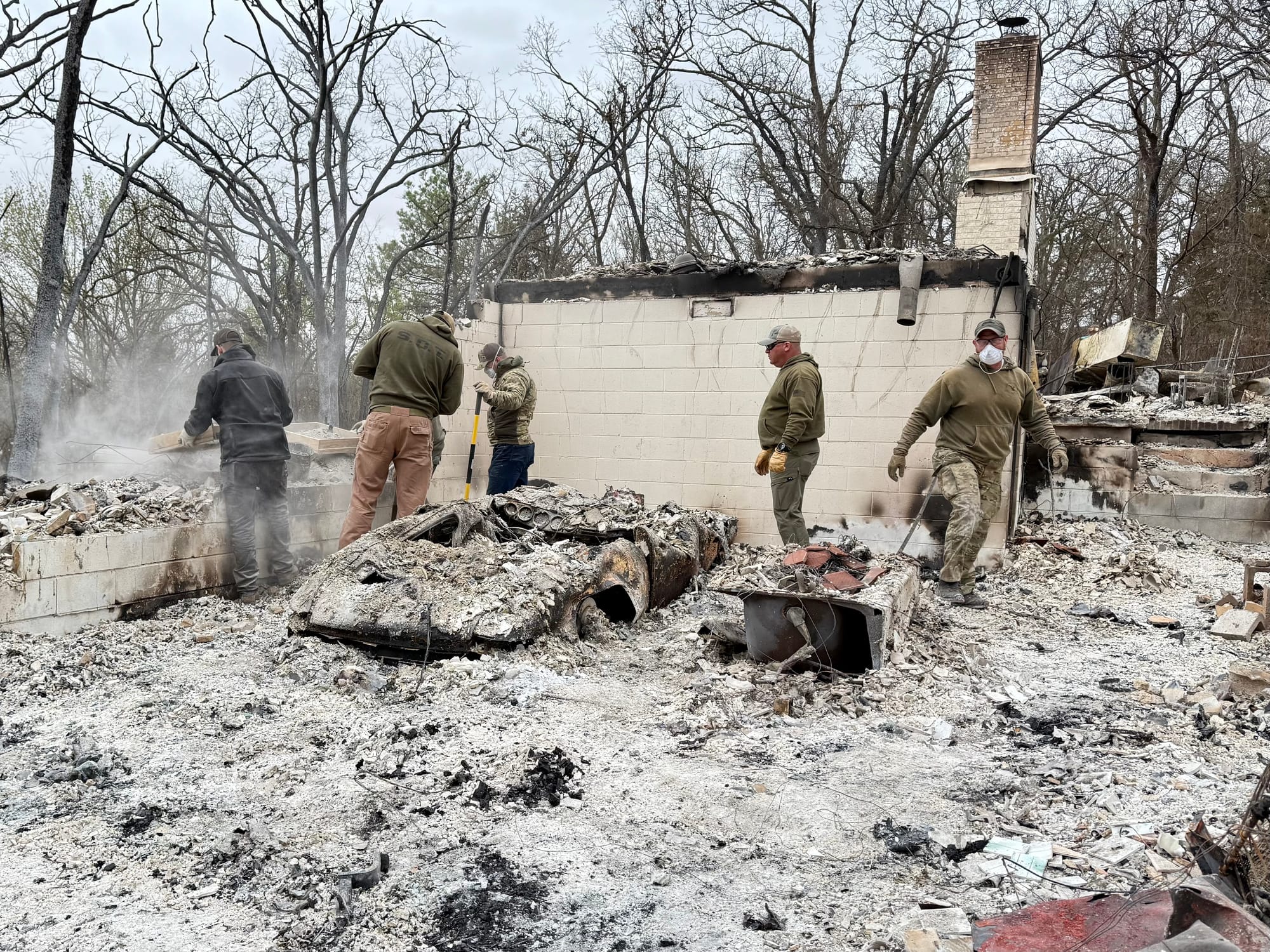 Search and rescue team members sift through ash-covered debris at a burned home site. A destroyed car frame lies in the foreground with a bathtub visible in the rubble. A partial concrete wall remains standing.
