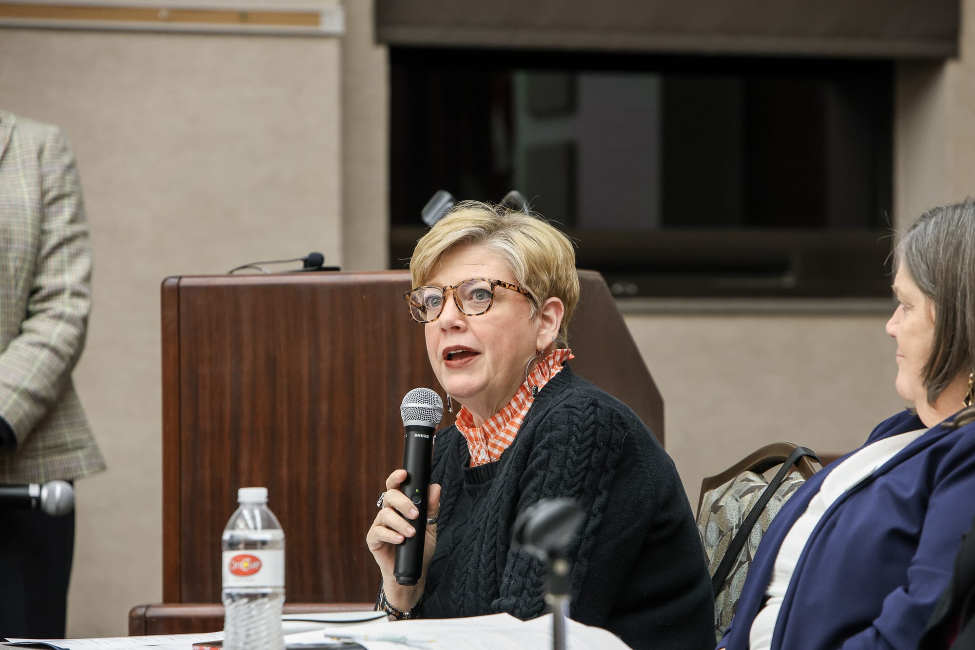 Brandi White addresses audience during panel discussion, holding microphone while seated at table with podium visible in background.