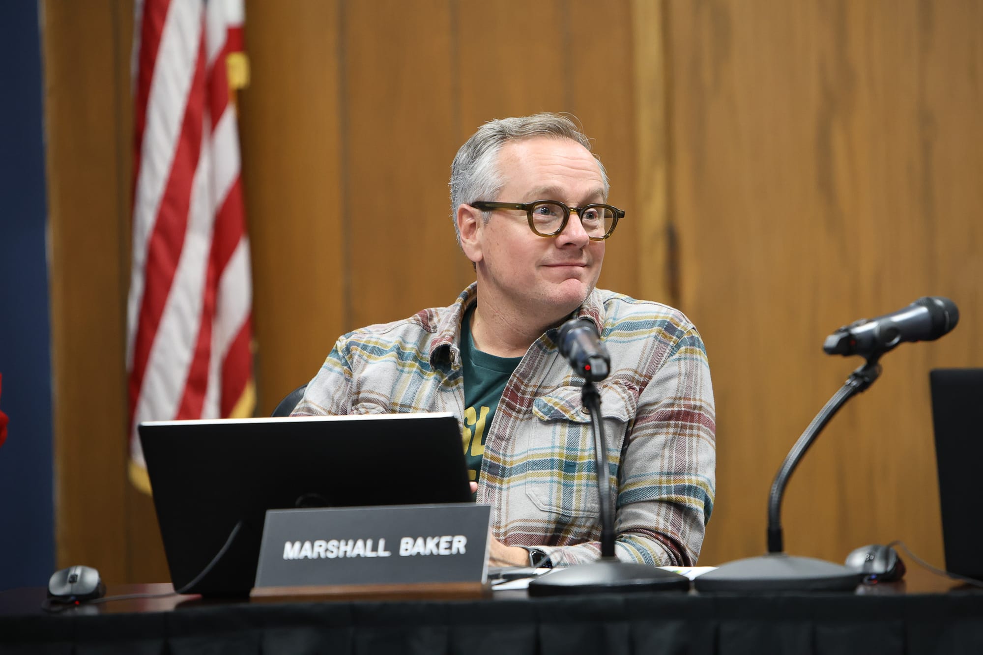 Dr. Marshall Baker sitting at board meeting table with laptop and microphone, American flag in background