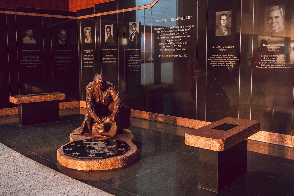 Remember the Ten Memorial in Gallagher-Iba Arena features bronze kneeling figure statue surrounded by memorial plaques with photos and tributes to 10 plane crash victims on dark walls.