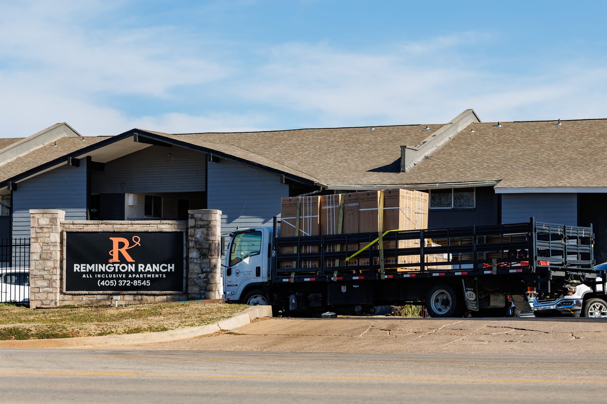 Delivery truck loaded with stacked water heater boxes parked at the entrance of Remington Ranch apartments, with the complex sign visible and apartment buildings in the background