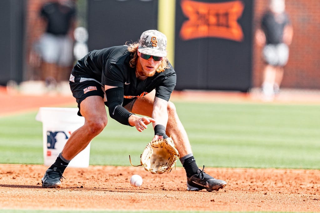 Oklahoma State baseball player Aidan Meola in black uniform fields ground ball during practice at O'Brate Stadium, bending low with glove positioned to scoop baseball on infield dirt