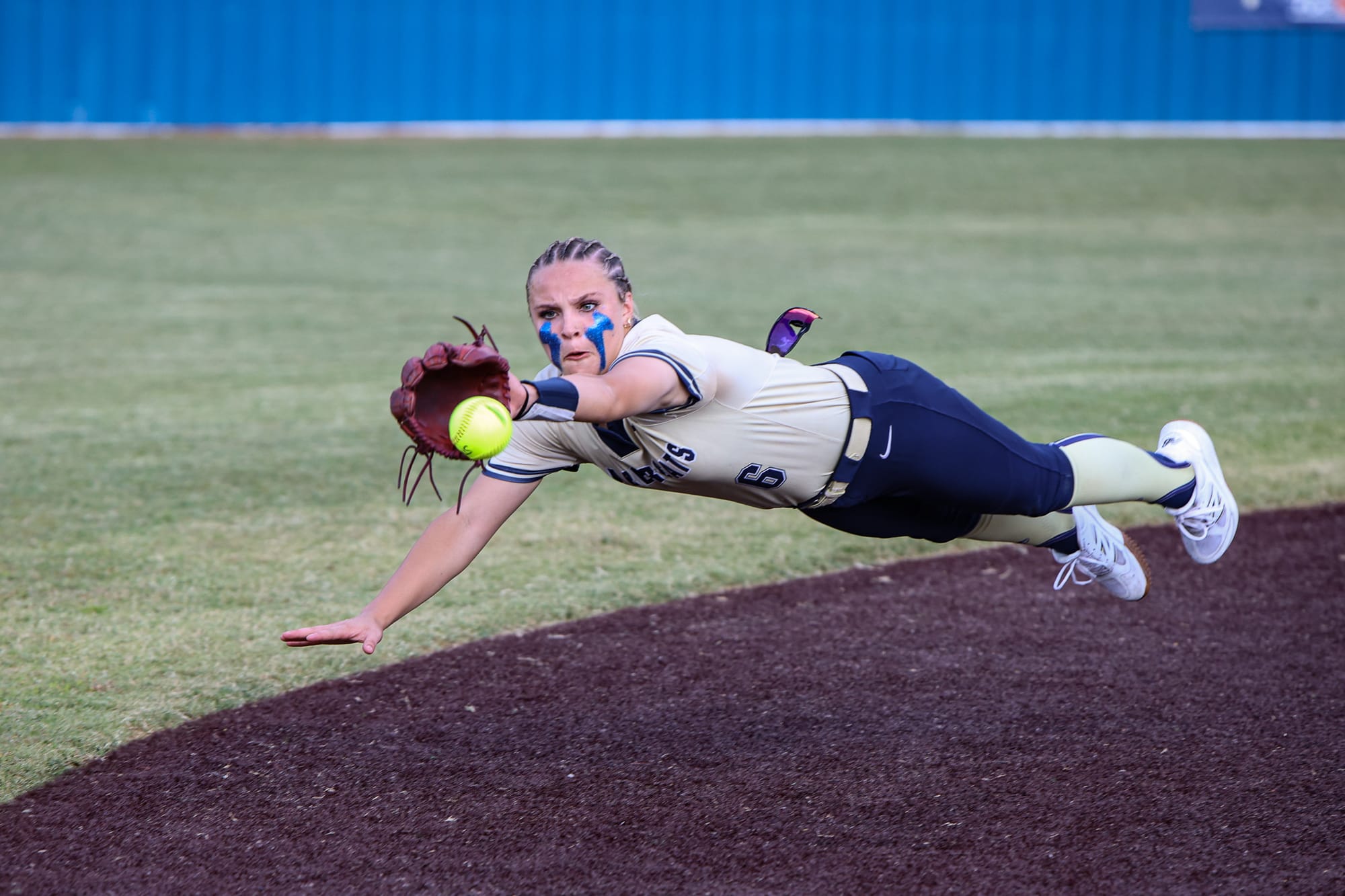 Softball player in gold and navy uniform diving horizontally through the air with glove extended toward yellow softball on infield dirt