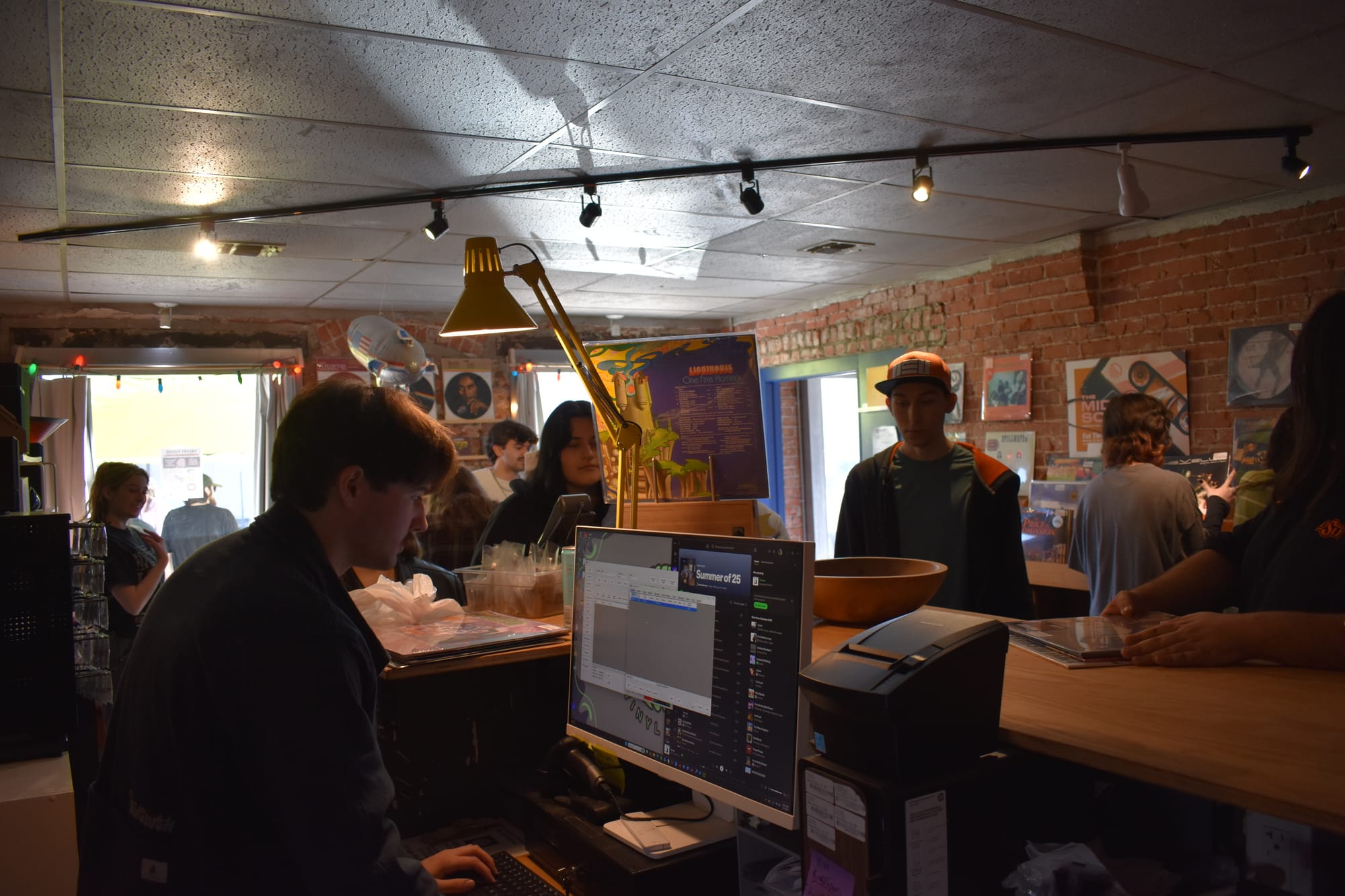 Store owner at checkout counter with computer monitor and register. Customers browse in background. Exposed brick walls, yellow desk lamp, track lighting, album posters visible.