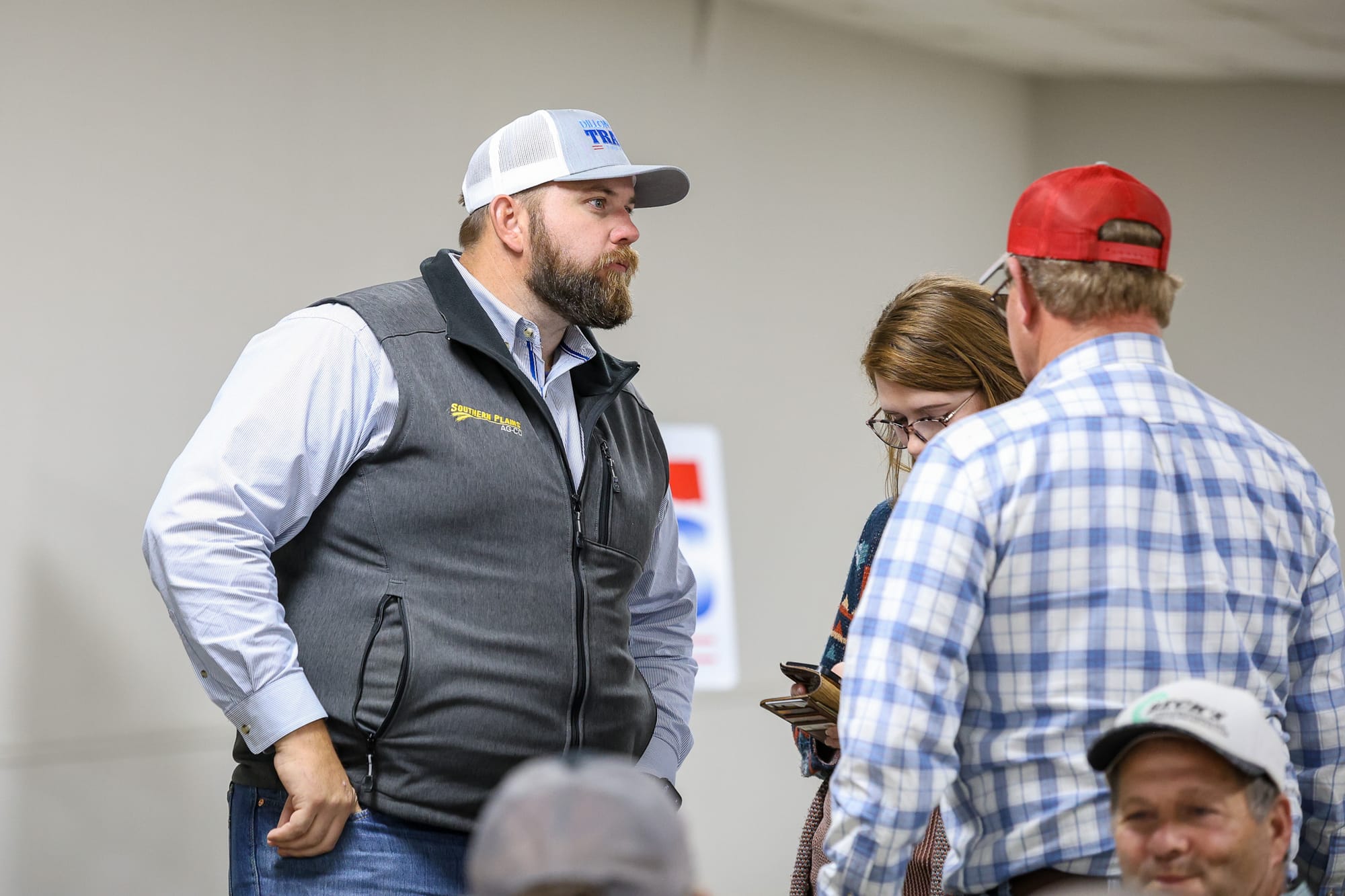 Dillon Travis wearing white trucker cap and dark Southern Plains vest over light blue shirt converses with group of attendees at indoor campaign event, with person in red cap and person in blue plaid shirt visible.