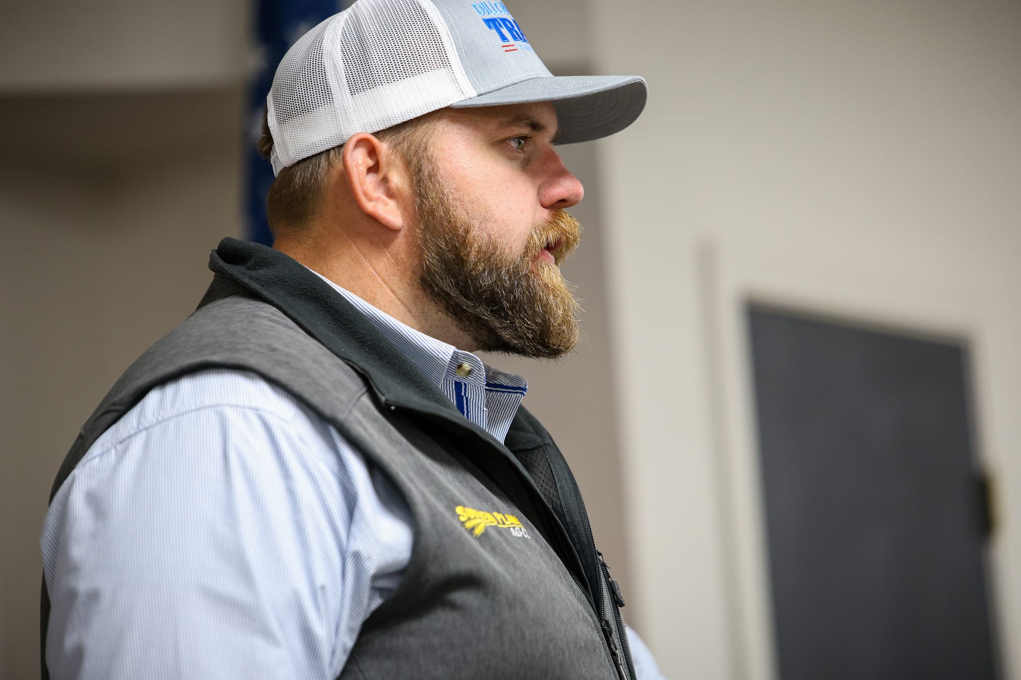 Profile view of Dillon Travis wearing trucker cap with Travis campaign logo and dark vest with Southern Plains branding over light blue striped shirt at indoor campaign event.