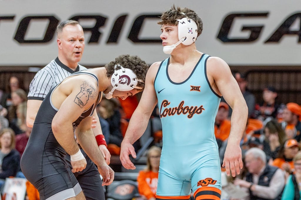 OSU wrestler Landon Robideau in sky-blue uniform raises his arm after pinning opponent at Gallagher-Iba Arena, with referee and Little Rock wrestler visible in background