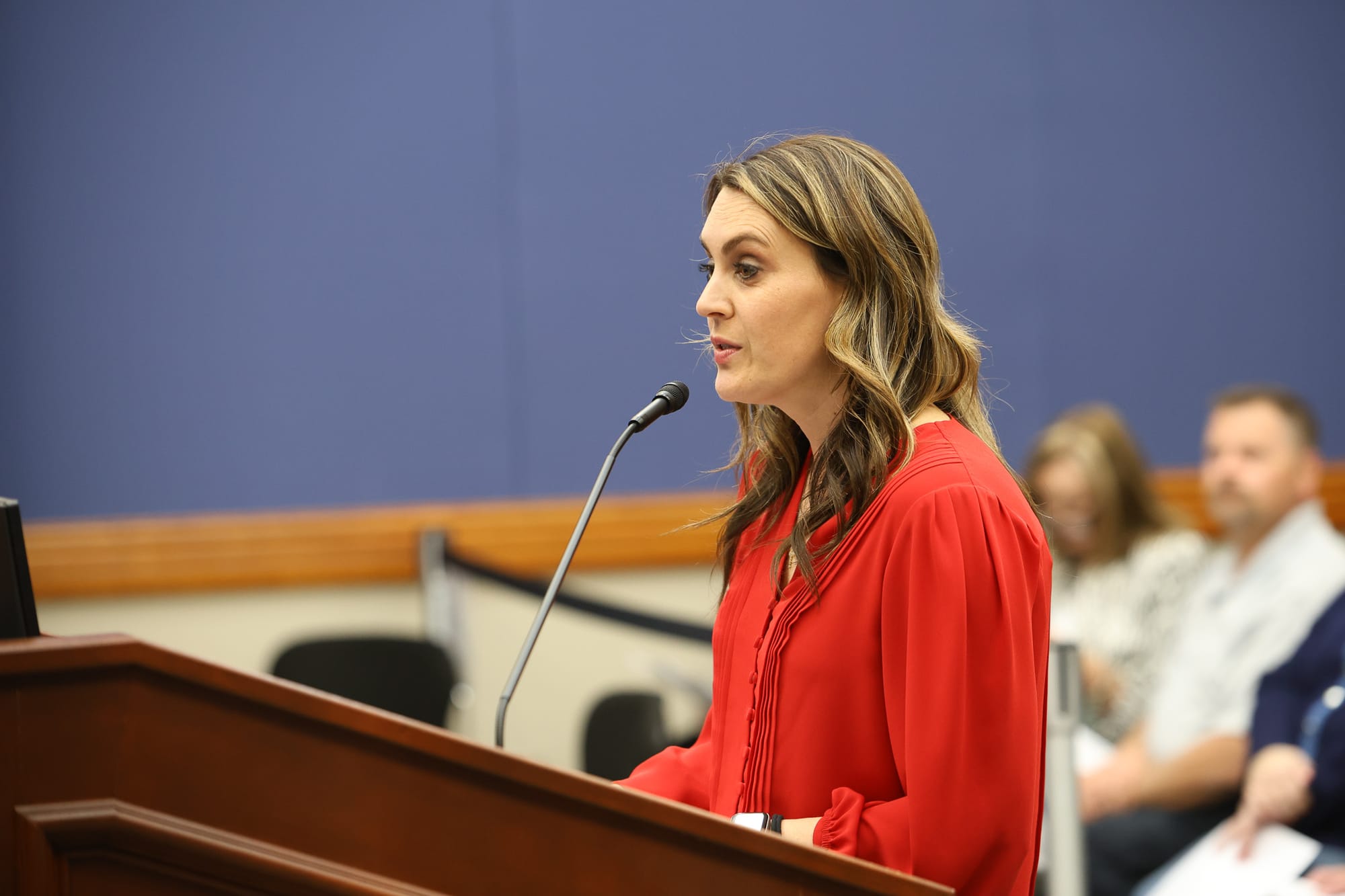 Laura Aufleger in red blouse speaks at podium with microphone during Stillwater City Council meeting to announce OnCue's $100,000 public art donation