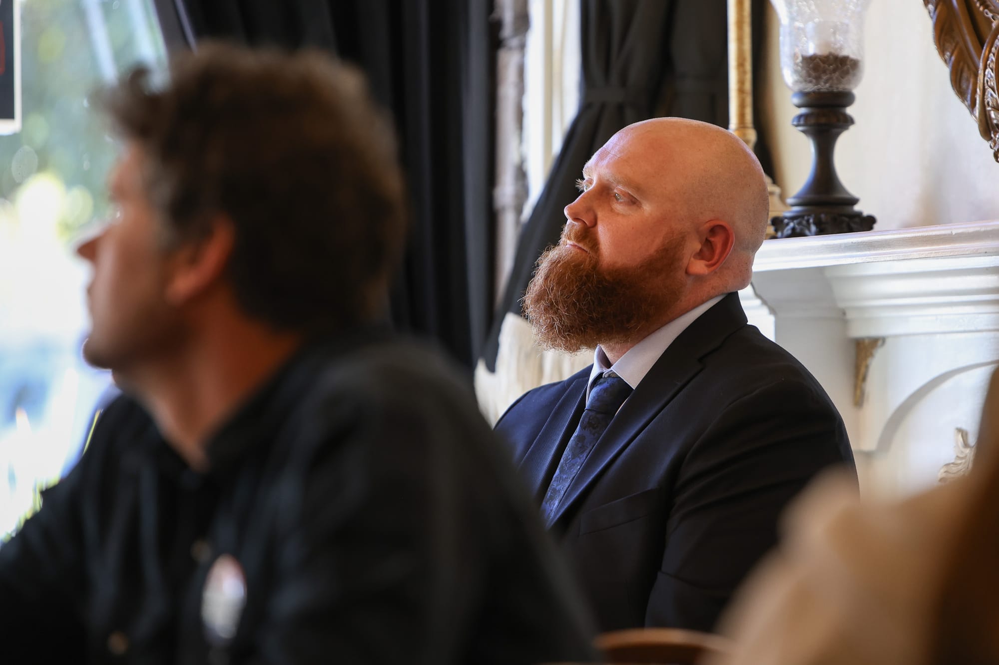 Luke Kruse wearing dark suit and blue tie sits looking upward while listening at indoor campaign event, with another attendee visible but blurred in foreground, dark curtains and white decorative column in background.