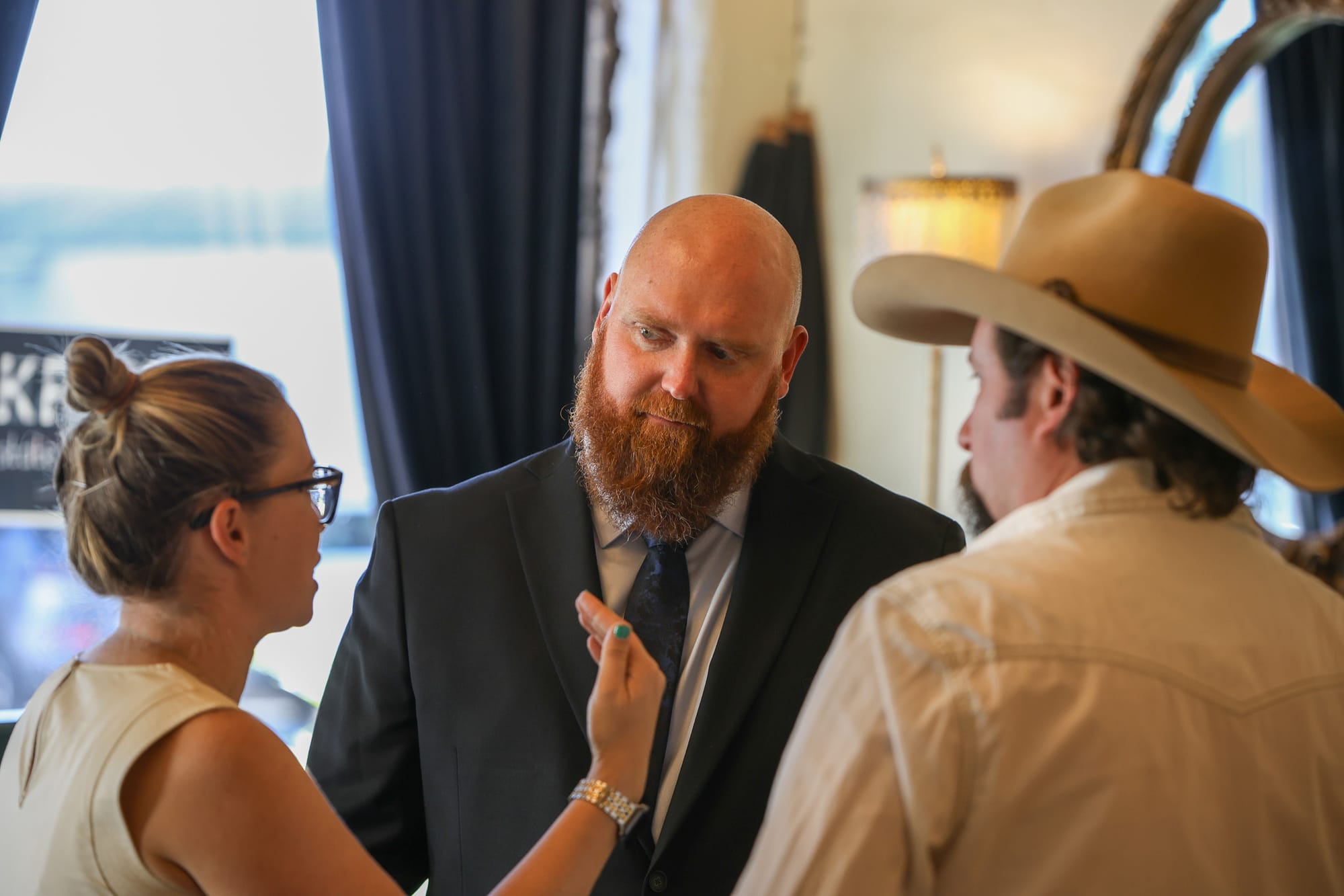Luke Kruse, man with reddish beard wearing dark suit and tie, speaks with two campaign event attendees in close conversation, with woman in glasses and sleeveless light top visible on left and person wearing tan cowboy hat visible on right, campaign signage and blue curtains in background.