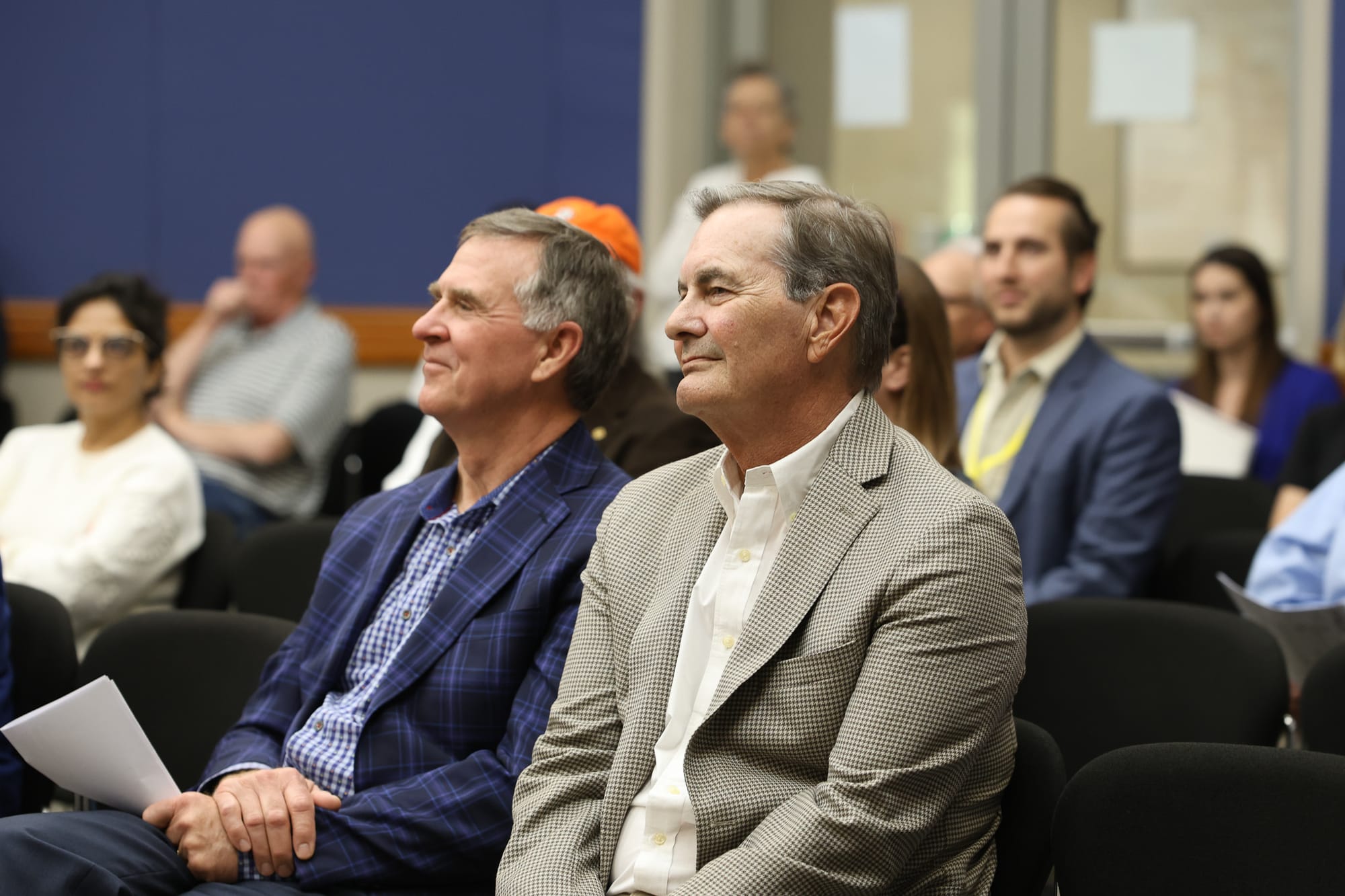 Two men in business attire seated in audience at Stillwater City Council meeting, other attendees visible in background