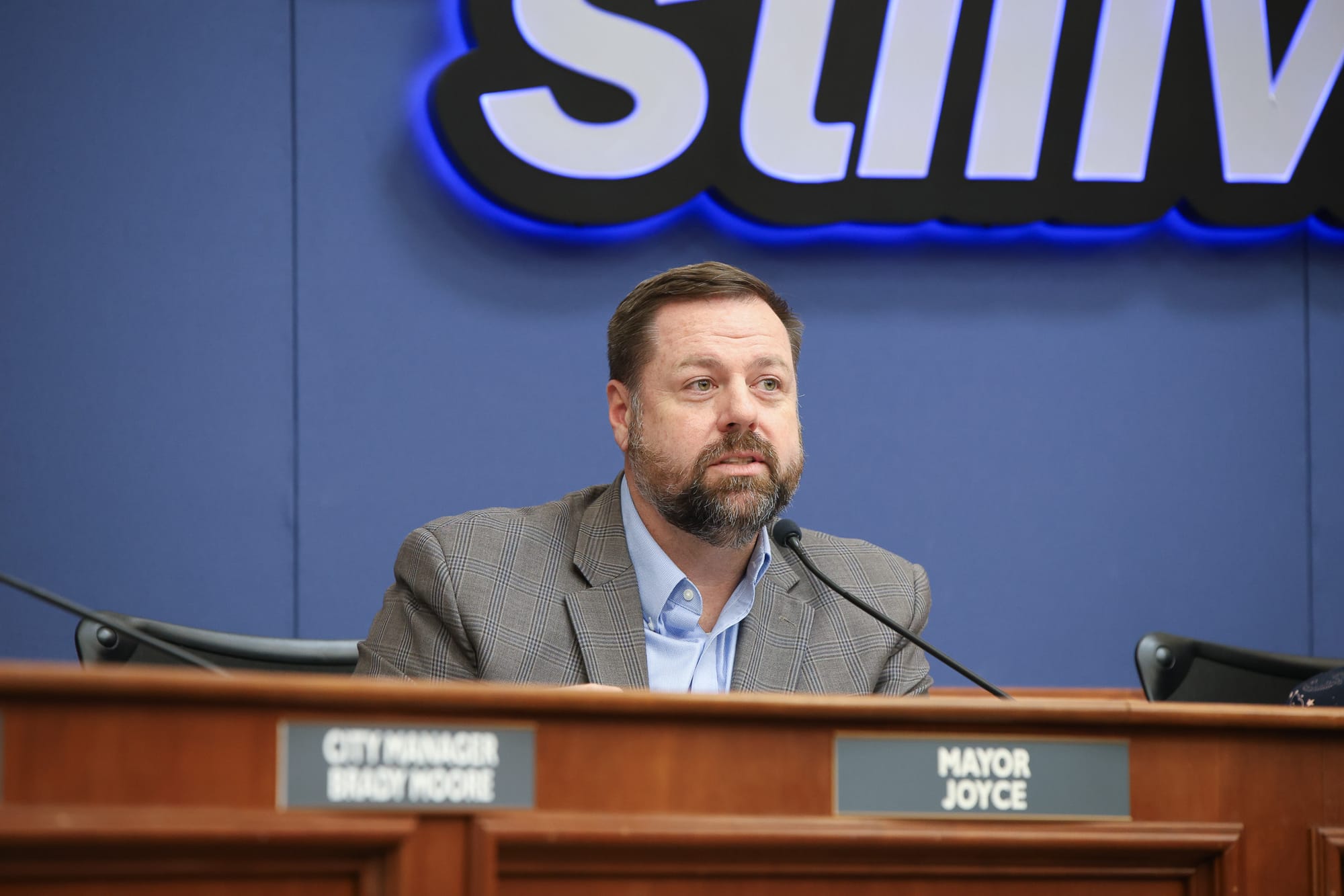 Stillwater Mayor Will Joyce sits at council dais with nameplate and microphone in front of him during city council meeting with blue Stillwater logo visible on wall behind him