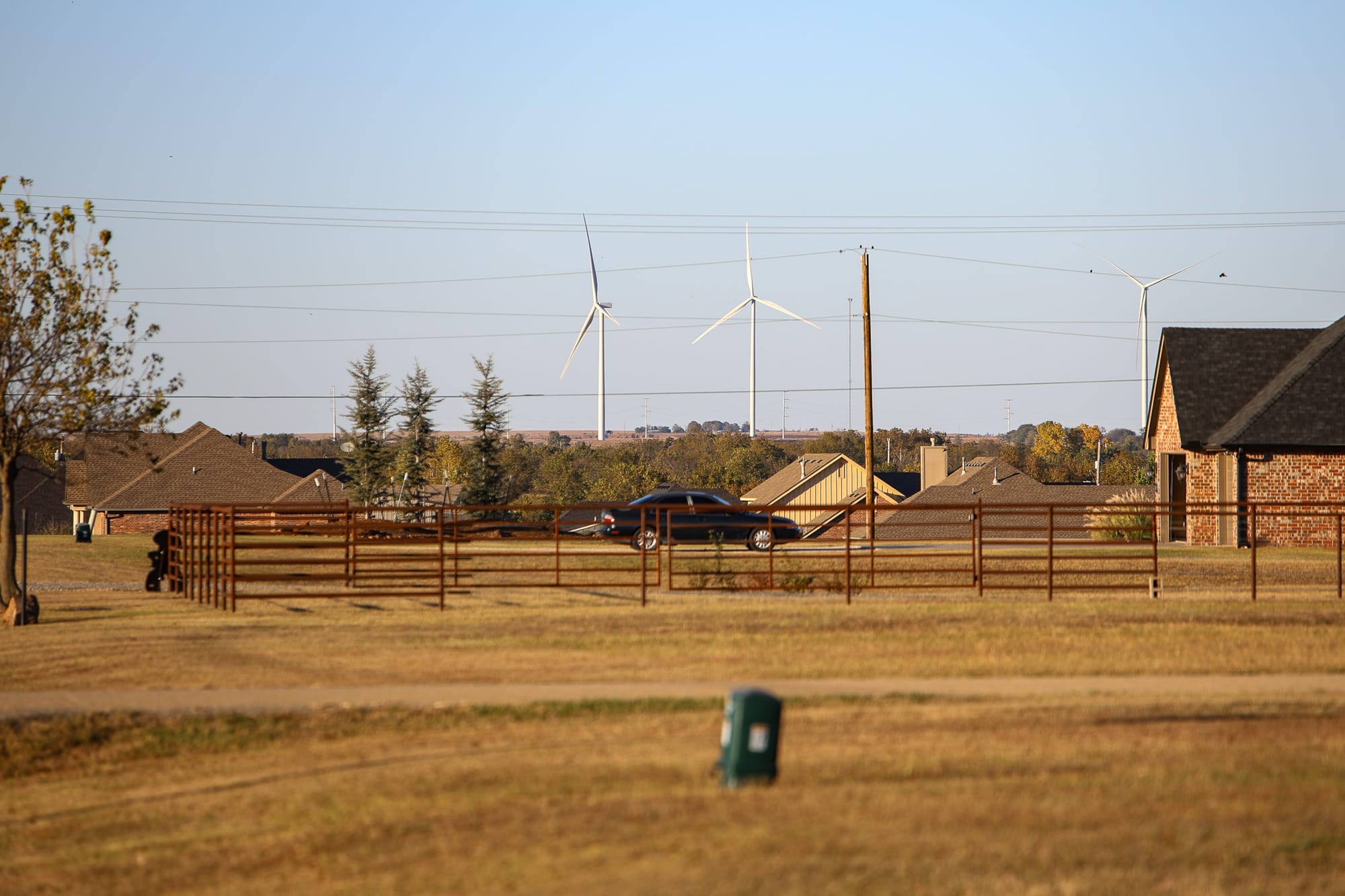 Landscape view of Morrison residential area with three tall white wind turbines visible in distance beyond suburban homes with brick exteriors and brown roofs, wooden ranch fencing crossing foreground, utility poles and power lines, and mix of green and brown grass under blue-gray sky.