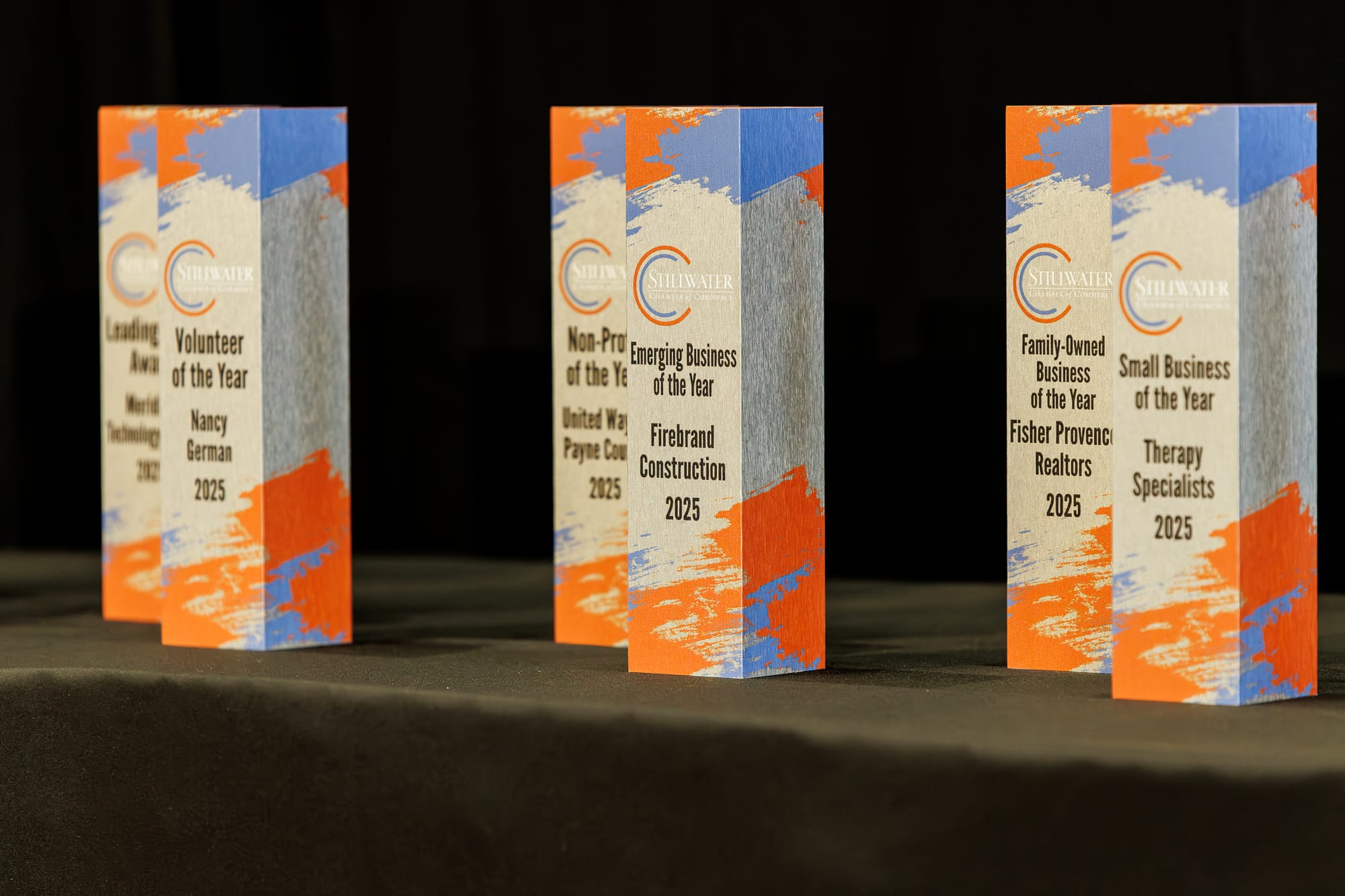 Six Stillwater Chamber of Commerce award trophies with orange, blue, and gray paint strokes stand on a table against a black backdrop before the Awards Gala ceremony.