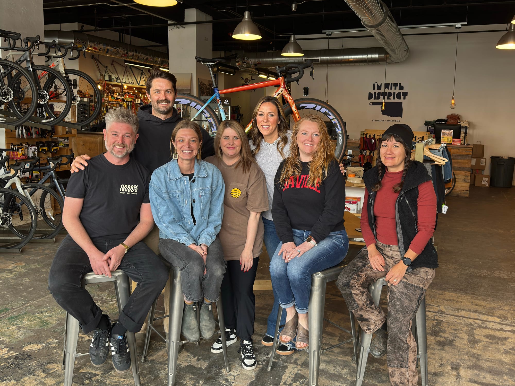 Seven people pose on stools inside District Bicycles in Stillwater with a custom-painted orange and red gravel bike displayed on a rack behind them. 