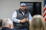 Dillon Travis wearing white baseball cap and gray Southern Plains Ag Co vest speaks to audience at indoor campaign event with American flag visible in background.
