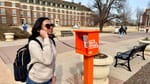 Student in sunglasses and backpack talks on orange OSU phone booth labeled "The Code Calls" on brick campus walkway with students passing in background