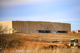 Large concrete tilt-up walls of Google's data center under construction, supported by black steel bracing, with excavators and bulldozers working on the graded dirt site under a blue sky.