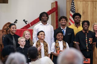 Nine youth speakers of varying ages pose together at Mt. Zion Baptist Church during Stillwater's MLK Jr. Day celebration, with American flag and red cross visible in background
