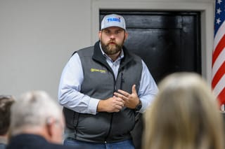 Dillon Travis wearing white baseball cap and gray Southern Plains Ag Co vest speaks to audience at indoor campaign event with American flag visible in background.