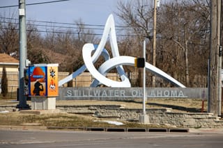 White metal sculpture called Interlace rises above stone wall reading "Stillwater Oklahoma" at intersection of Sixth Avenue and Western Road, with decorated utility box visible at left.