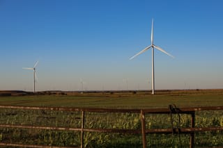 Wind turbines rise above green agricultural fields with a rusty barbed wire fence in the foreground under a clear blue sky in Noble County, Oklahoma