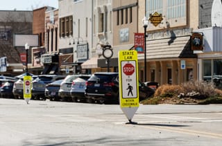 Yellow pedestrian crossing signs reading "State Law: Stop for pedestrians within crosswalk" stand at the intersection of South Main Street and Seventh Avenue in downtown Stillwater.