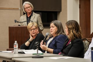 Janet Fultz speaks into a microphone during a panel discussion at Stillwater Public Library, with other panelists seated at the table and a moderator standing in the background.