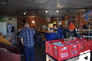 Record store interior with customers browsing vinyl in red crates in foreground. Owner at checkout counter in background. Brick walls with album posters, track lighting.