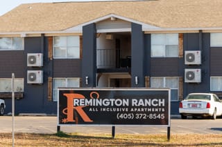 Sign reading "Remington Ranch All Inclusive Apartments" stands before weathered gray apartment building with AC units mounted on exterior walls.