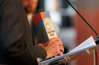 Steve Irby holds a wooden Hall of Fame Award trophy with red, blue, and gray paint strokes at a clear podium during the Stillwater Chamber Awards Gala.