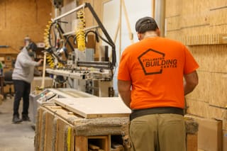 Two Stillwater Building Center employees work in the company's custom door shop, one inspecting a door panel, another operating fabrication machinery in the background.