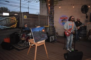 Billy Bonney performs on the Stonecloud Brewing patio. The As The Canyons Roar album cover painting sits on an easel in the foreground; the band's banner hangs behind the drum kit.