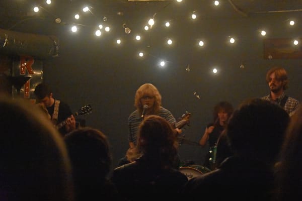 Band EZ4U2SEY performs on stage at The Bait Shop venue with string lights overhead and audience members visible in the foreground.