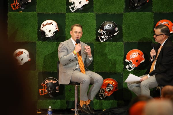 Eric Morris in gray suit with orange tie speaks into microphone during Oklahoma State football coach introduction, with OSU helmets displayed on wall behind him