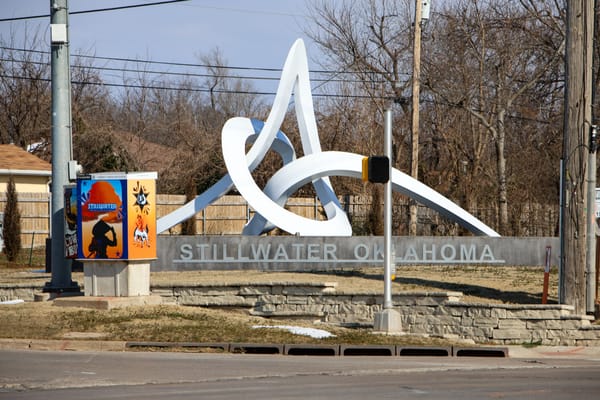 White metal sculpture called Interlace rises above stone wall reading "Stillwater Oklahoma" at intersection of Sixth Avenue and Western Road, with decorated utility box visible at left.
