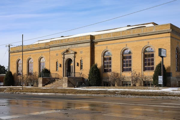 Historic tan brick neoclassical library building with "PUBLIC LIBRARY" text on facade, featuring arched windows, central entrance with pediment, and curved staircases.