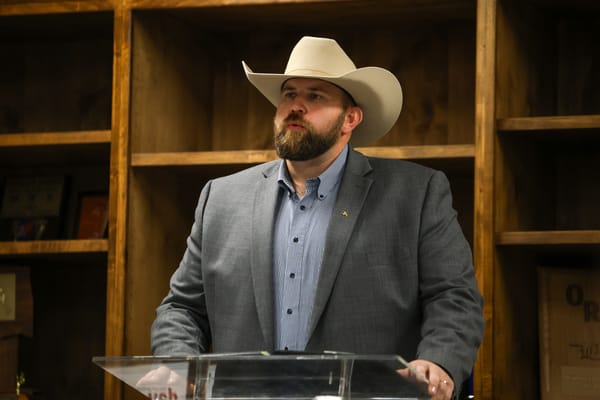 Dillon Travis wearing tan cowboy hat and gray suit jacket stands at clear podium while speaking at indoor campaign event with wooden cabinetry visible in background.