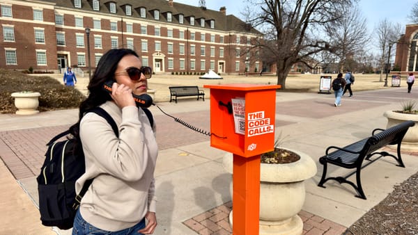 Student in sunglasses and backpack talks on orange OSU phone booth labeled "The Code Calls" on brick campus walkway with students passing in background