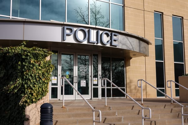 Front entrance of Stillwater Police Department building with tan brick facade, curved POLICE awning over glass doors, metal handrails, and concrete steps