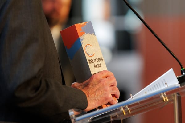 Steve Irby holds a wooden Hall of Fame Award trophy with red, blue, and gray paint strokes at a clear podium during the Stillwater Chamber Awards Gala.