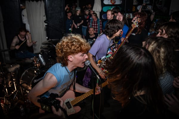 A curly-haired guitarist leans toward the crowd, singing into a microphone at a packed indoor venue. 