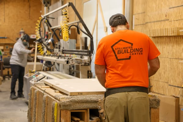 Two Stillwater Building Center employees work in the company's custom door shop, one inspecting a door panel, another operating fabrication machinery in the background.