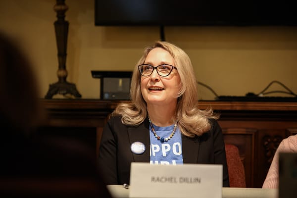 Rachel Dillin, wearing a black blazer and campaign button, sits at a table during a candidate forum. A nameplate reading "Rachel Dillin" is visible in the foreground.